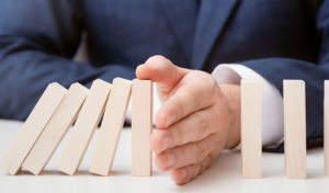 foto de uma pessoa que usa terno e gravata e que interrompe com uma das mãos, que está sobre a mesa, a queda de peças enfileiradas.